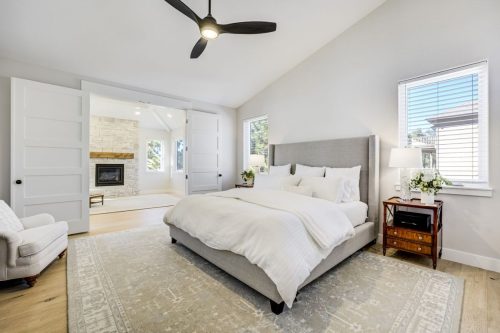 Primary bedroom sitting area with stacked stone gas fireplace, wood mantel, and upholstered armchair with ottoman